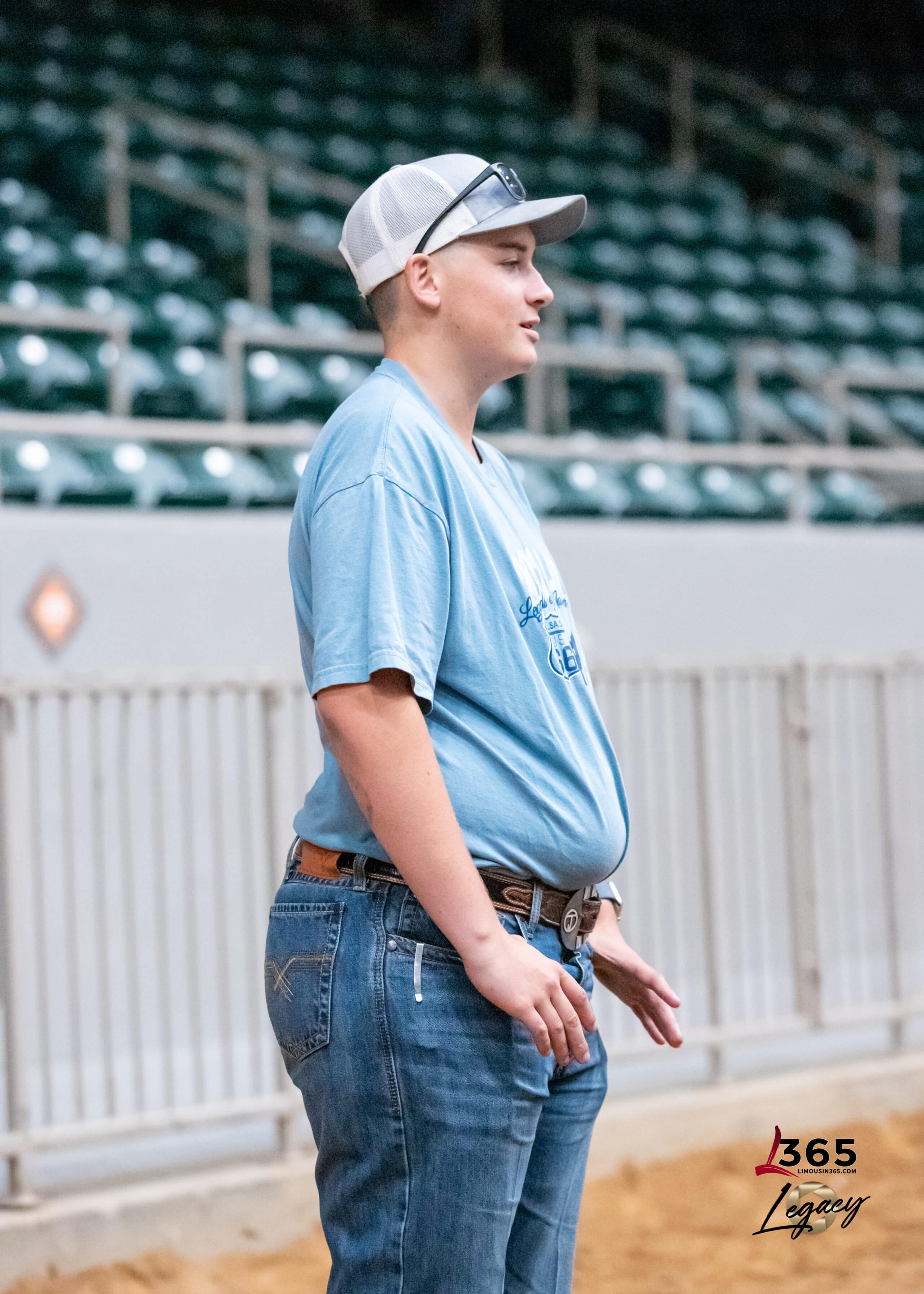 A young person in a blue T-shirt, jeans, and a white cap stands indoors near metal railings and empty bleachers, looking to the side. The floor is sandy, and there is a 365 Legacy watermark in the corner.