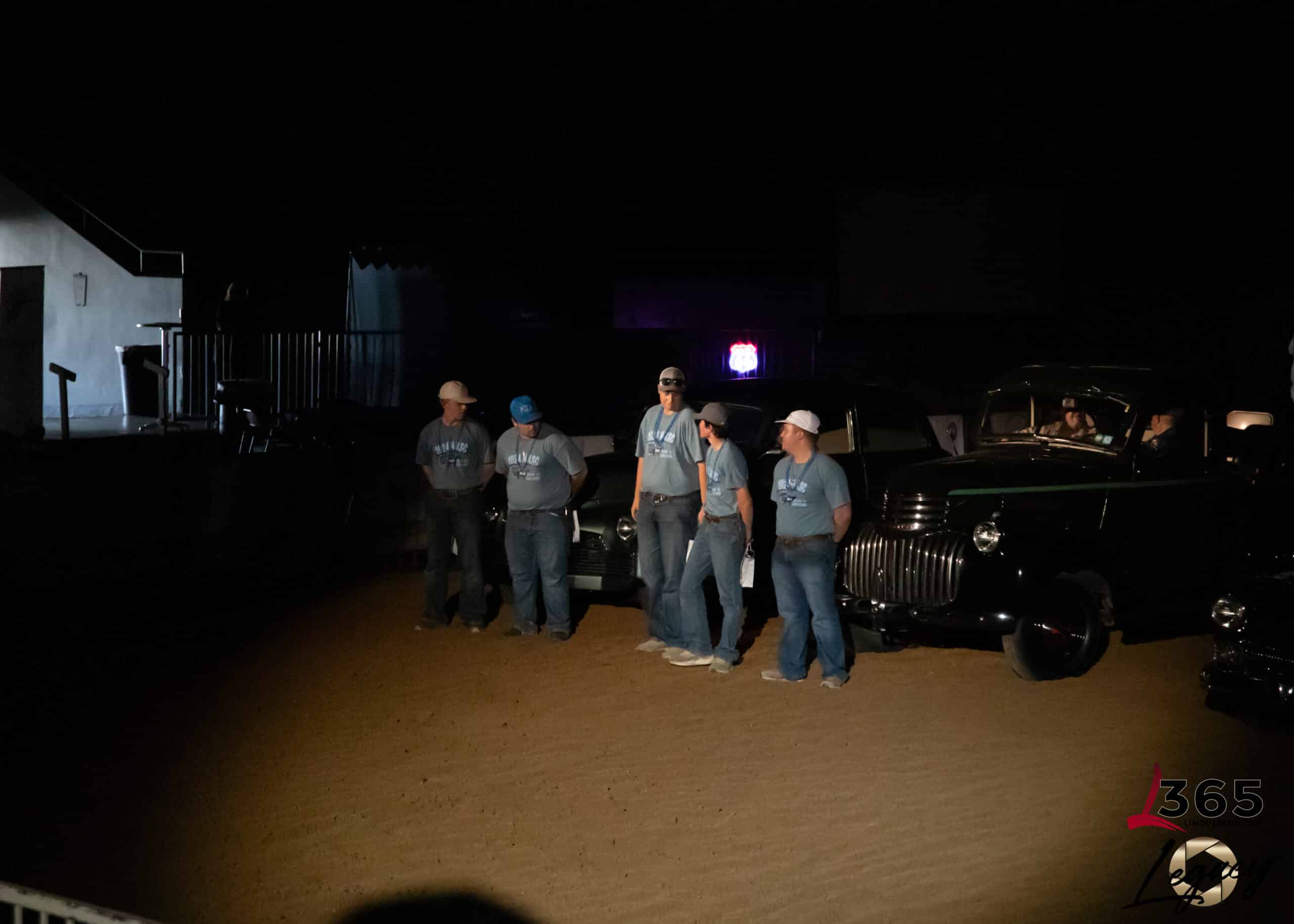 Five people in matching shirts and hats stand in a row on a dirt surface at night, illuminated by a spotlight, with classic cars partially visible behind them and darkness surrounding the scene.