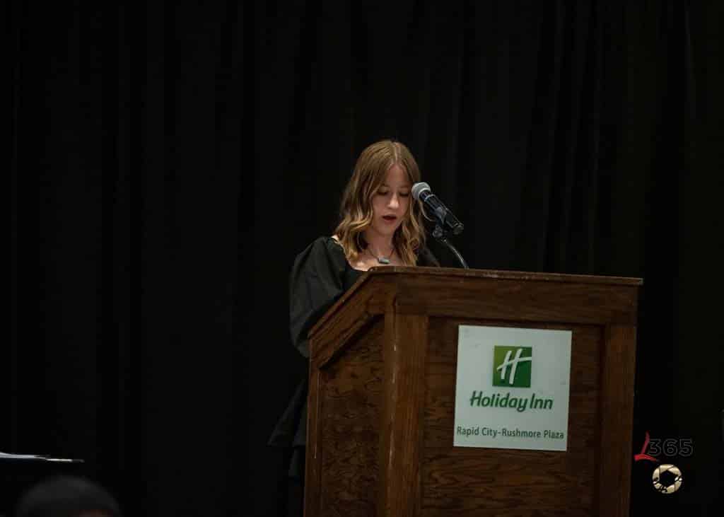 A woman with shoulder-length hair speaks at a podium with a microphone. The podium displays a Holiday Inn sign. The background is dark, and the setting appears to be an event or conference.