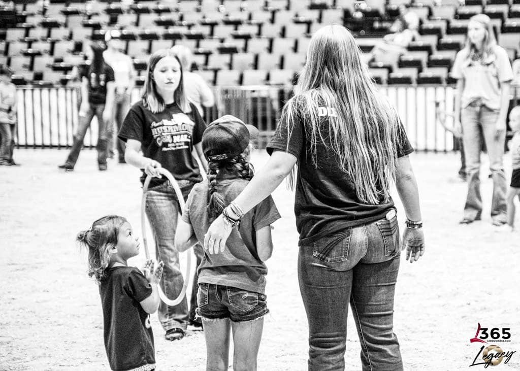Four girls stand in a sandy arena, with one adult guiding three children. Empty seats fill the background and a logo reading “365 Legacy” appears in the lower right corner. The image is black and white.