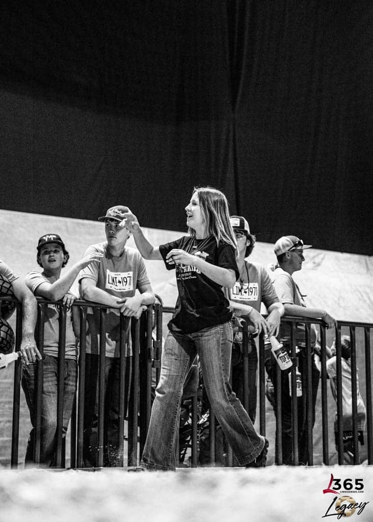 A woman stands by a railing, gesturing with her right hand, while several young people in hats and numbered shirts watch her. The scene appears to be at an indoor event. The photo is in black and white.