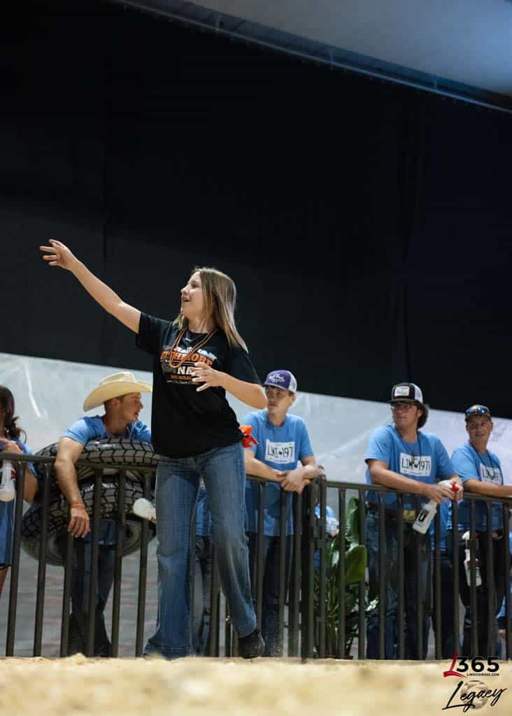 A woman in a black shirt and jeans throws an object while several people in blue shirts watch from behind a fence at an indoor event. The floor is covered in straw, and the background is dark.