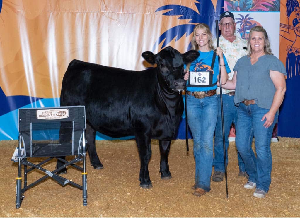 Four people stand with a black cow at a livestock show; one young woman holds the cow’s lead and a plaque marked 162. A branded chair and a colorful backdrop are visible behind them.