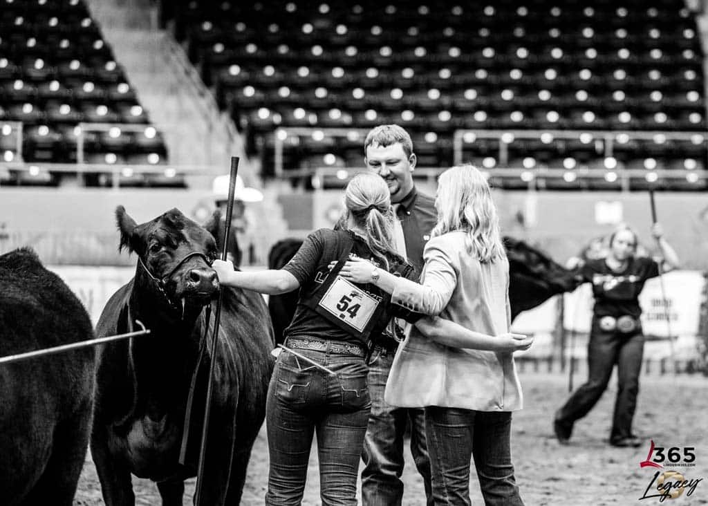 A black and white photo of three people standing by a cow at a livestock show, sharing a hug. Another person leads a cow in the background. The arena seats are mostly empty. The number 54 is visible on one persons back.