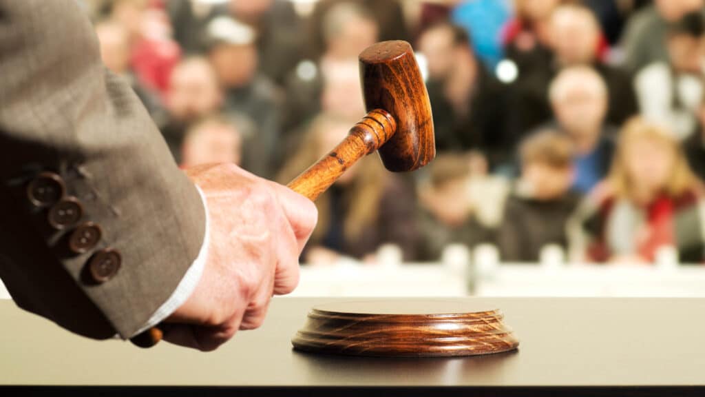A close-up of a judge’s hand holding a wooden gavel above a sound block, with a blurred crowd of people in the background.