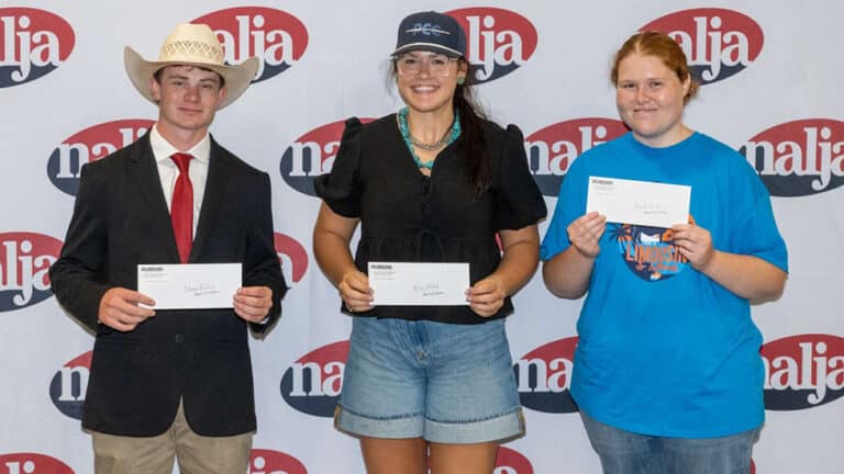 Three young people stand side by side holding envelopes, posing in front of a backdrop with a repeating najja logo. The person on the left wears a cowboy hat and suit, the middle person wears glasses and shorts, and the right person wears a blue shirt.