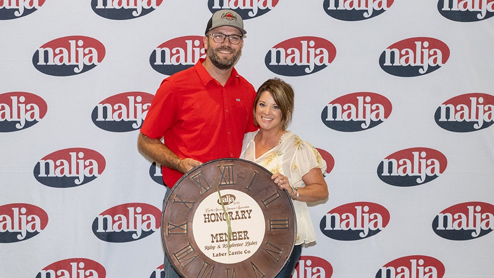 Two people stand smiling in front of a backdrop with repeated nalja logos. They hold a large clock-shaped award that reads Honorary Member along with the nalja logo and other text.