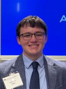 A young man in glasses, a suit, and a tie smiles at the camera. He is wearing a name tag, and the background is a bright blue screen.