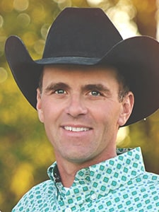 A smiling man wearing a black cowboy hat and a patterned button-up shirt stands outdoors with a blurred background of green and yellow foliage.