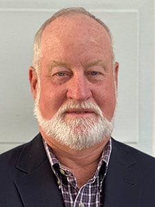 A middle-aged man with a white beard and short hair, wearing a dark blazer over a checkered shirt, poses in front of a light-colored background.