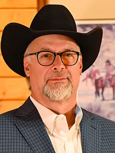 A man with a gray beard and glasses, wearing a black cowboy hat, white shirt, and blue checked blazer, stands indoors with a wooden wall and a painting in the background.