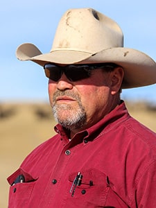 A man wearing a beige cowboy hat, dark sunglasses, and a red button-up shirt stands outdoors in a sunny, rural setting. He has a serious expression and pens in his shirt pocket.