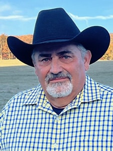 A man with a gray beard wearing a black cowboy hat and a yellow and blue checkered shirt stands outdoors in a field with trees and a blue sky in the background.