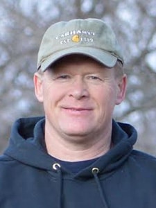 A man wearing a green Carhartt cap and a dark hoodie smiles at the camera. The background is outdoors with blurred trees.