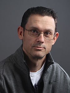 A man with short dark hair, glasses, and a trimmed beard wears a gray zip-up jacket over a white shirt, looking at the camera with a neutral expression against a plain gray background.