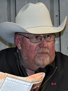 A man wearing a white cowboy hat and glasses is holding a magazine or brochure. He has a gray beard and is dressed in a dark jacket with Rob embroidered on it. The background appears to be an indoor setting.