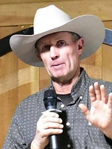 A man wearing a light-colored cowboy hat and patterned shirt holds a microphone and gestures with his hand while speaking, standing in front of a wooden background.
