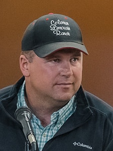 A man wearing a black Coleman Limousin Ranch cap and a dark Columbia jacket speaks into a microphone. He is facing forward with a neutral expression against a brownish background.