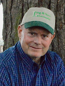 A man wearing a green and beige cap and a blue plaid shirt sits in front of a tree, smiling gently at the camera. The cap has green writing that reads “Long & Sons Limousin.”.