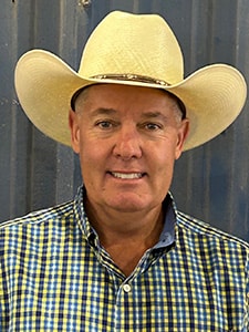 A man wearing a light-colored cowboy hat and a yellow and blue checkered shirt smiles at the camera; he stands in front of a corrugated metal wall.