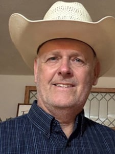 A middle-aged man wearing a light-colored cowboy hat and a dark plaid shirt smiles at the camera indoors, with a framed window and part of a picture visible in the background.