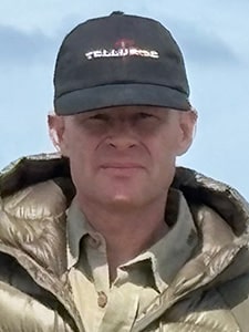 A person wearing a black Telluride cap, a beige collared shirt, and a shiny, light-colored puffer jacket is standing outdoors with a blue sky in the background.