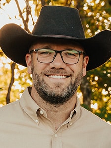 A smiling man with a beard and glasses wearing a black cowboy hat and a beige button-up shirt stands outdoors with sunlight and trees in the background.