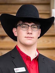 A young man wearing glasses, a black cowboy hat, a red shirt, and a dark blazer stands in front of wooden steps. He has a name badge pinned to his jacket.