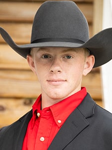 A young man wearing a large black cowboy hat, a black blazer, and a bright red collared shirt stands in front of a wooden background, looking directly at the camera with a neutral expression.
