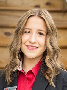 A young woman with wavy blonde hair smiles at the camera. She is wearing a black jacket over a red collared shirt, and stands in front of a wooden background.