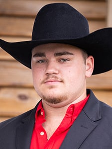 A young man wearing a black cowboy hat, a black blazer, and a red shirt stands in front of a wooden wall, looking at the camera with a neutral expression.