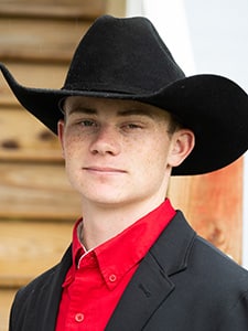 A young person with light skin, freckles, and short hair wears a black cowboy hat, red shirt, and black blazer. Wooden steps and a white railing appear in the blurred background.