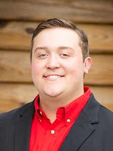 A man with short brown hair, wearing a red button-up shirt and a black blazer, smiles in front of a wooden wall background.