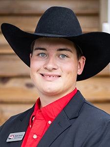 A young person wearing a black cowboy hat, a black blazer, and a red collared shirt smiles at the camera. The background features wooden planks.