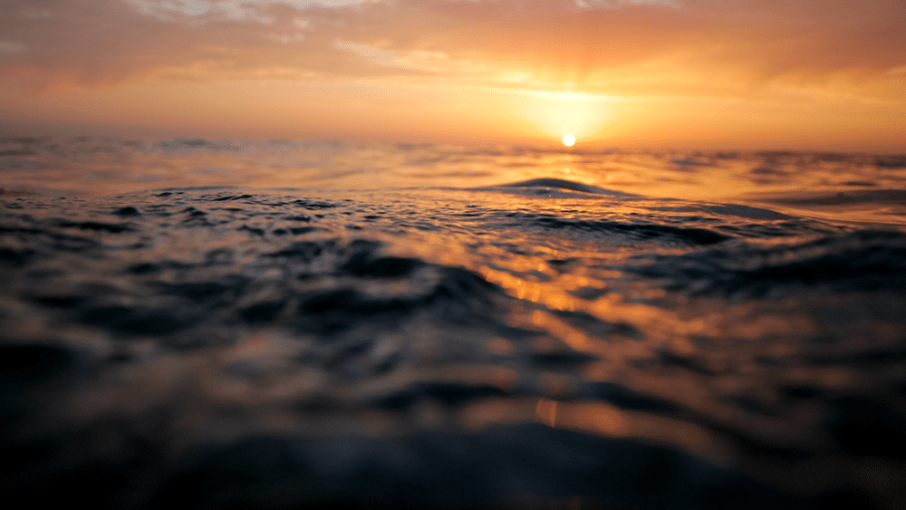 Close-up view of ocean waves with the sun setting on the horizon, casting warm orange and yellow hues across the sky and reflecting on the water’s surface.
