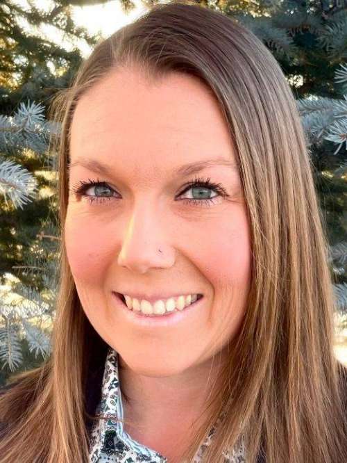 A woman with long straight hair and light eyes smiles outdoors in front of pine tree branches. She is wearing a patterned shirt and has a small nose piercing. Sunlight softly lights her face.