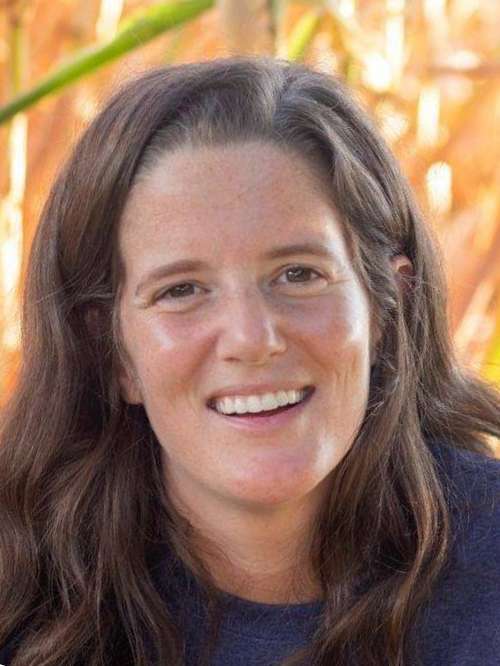 A woman with long brown hair and a friendly smile is outdoors, with tall, sunlit grass or plants in the blurred background. She is wearing a dark top.