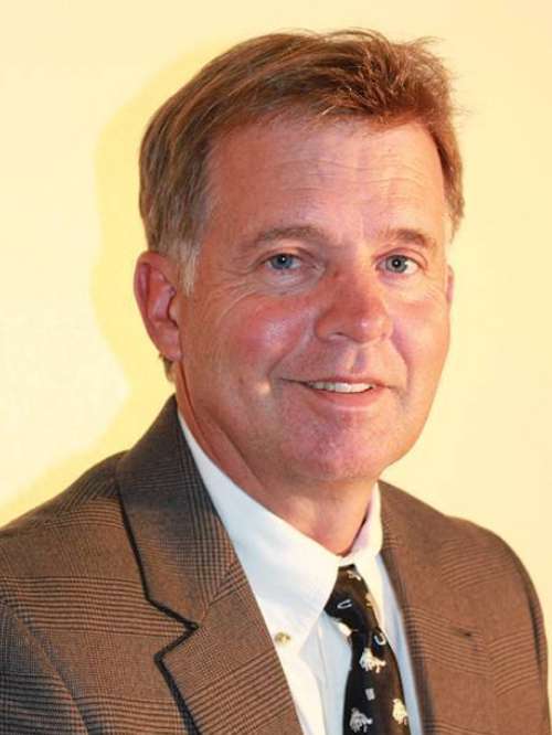 Middle-aged man with short light brown hair, wearing a brown plaid suit jacket, white shirt, and black tie with white patterns, smiling slightly against a pale yellow background.