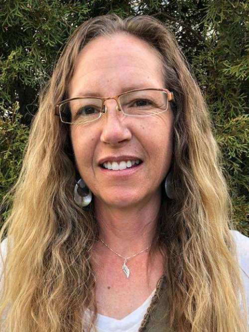 A woman with long, wavy blonde hair and glasses smiles outdoors. She is wearing a white top, silver earrings, and a necklace with a pendant. Green foliage is visible in the background.