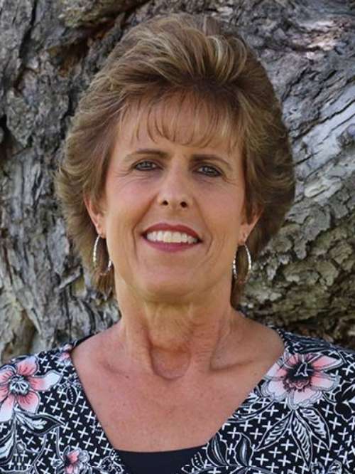A woman with short, light brown hair and hoop earrings smiles at the camera. She is wearing a black and white patterned top with pink flowers, standing in front of a textured tree trunk.