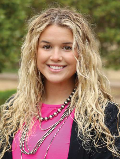 A woman with long, wavy blonde hair smiles at the camera. She is wearing a bright pink top, layered necklaces, and a black blazer. The background is blurred greenery.