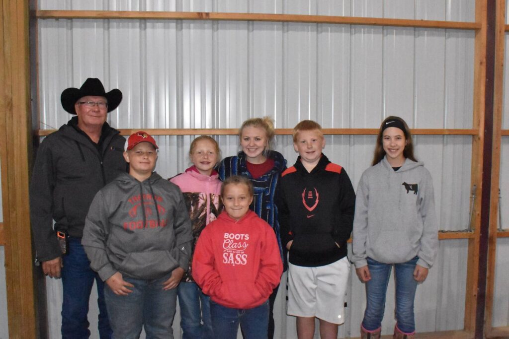 A man in a black cowboy hat stands with six smiling children in casual clothes and hoodies, posing in front of a corrugated metal wall inside a building.
