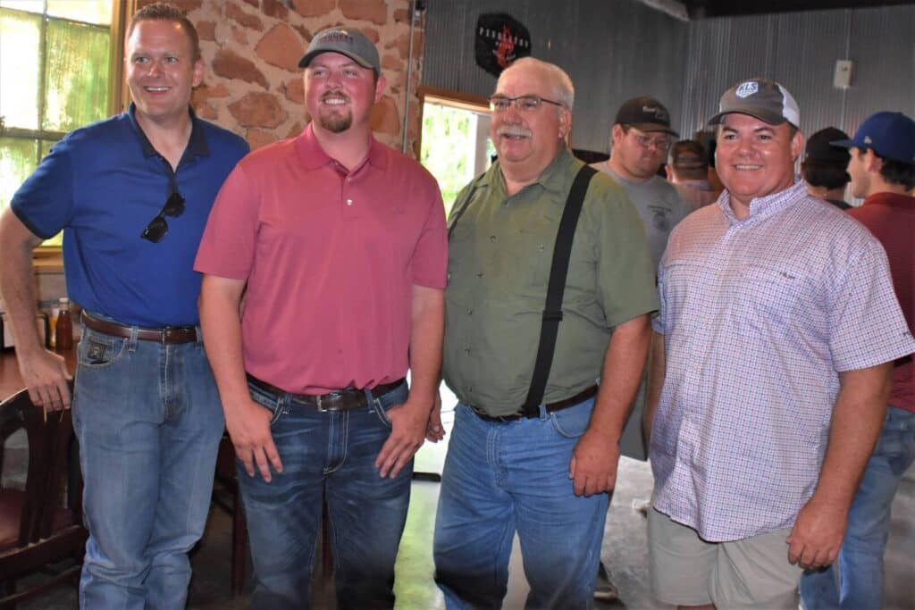 Four men stand side by side indoors, smiling at the camera; they are casually dressed in jeans, polo shirts, and button-ups, with other people and rustic decor visible in the background.
