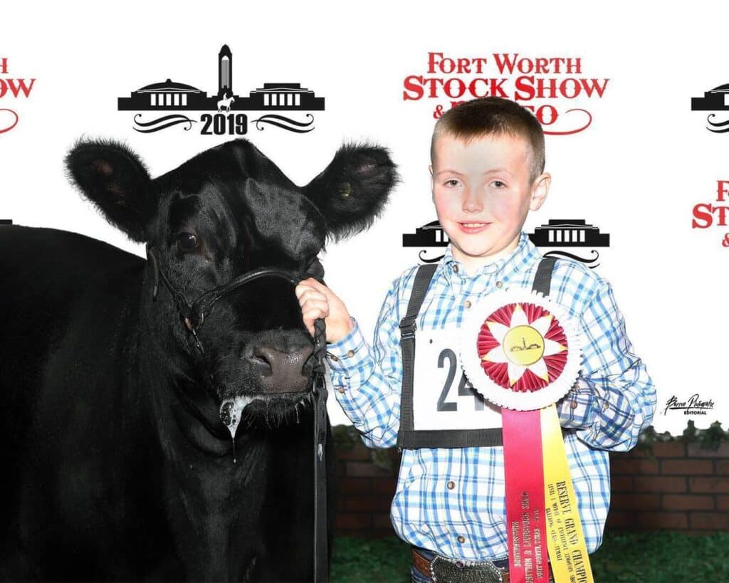 A boy stands smiling next to a black cow, holding its lead and a prize ribbon, in front of a Fort Worth Stock Show 2019 backdrop.