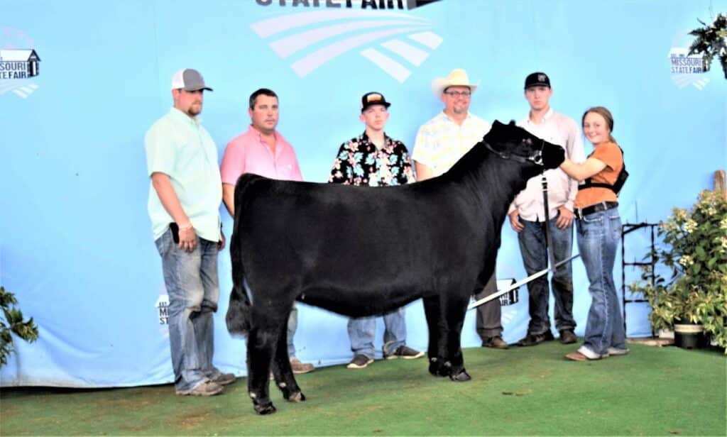 Six people stand behind a black steer at the Missouri State Fair, posing for a photo against a blue backdrop with the fair logo. One woman holds the steer by a halter.