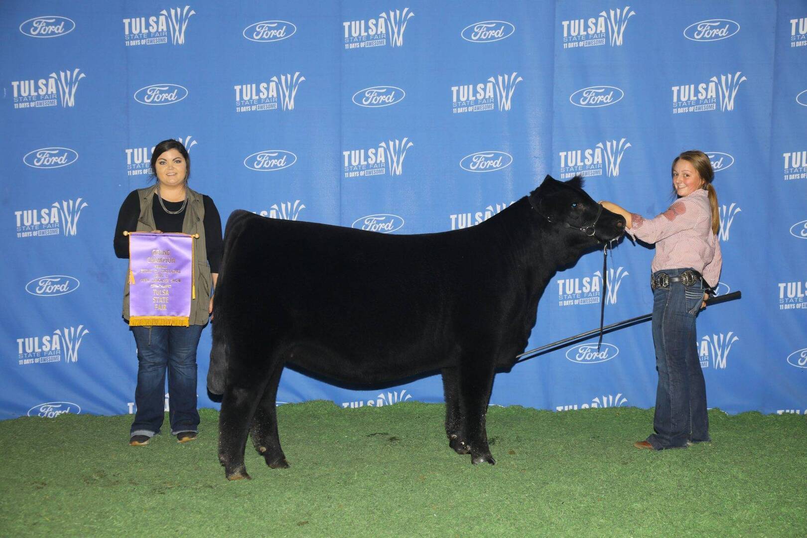 Two young women pose with a black cow in front of a blue backdrop with “Tulsa State Fair” and “Ford” logos; one woman holds a purple award banner, and the other holds the cow’s lead.