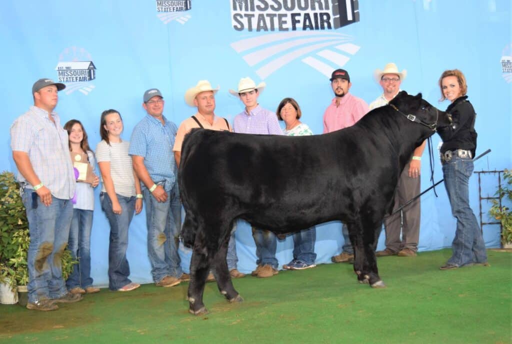 A group of people poses with a black cow in front of a blue Missouri State Fair backdrop. Some hold awards, and a woman in black stands at the cows head holding a lead rope.