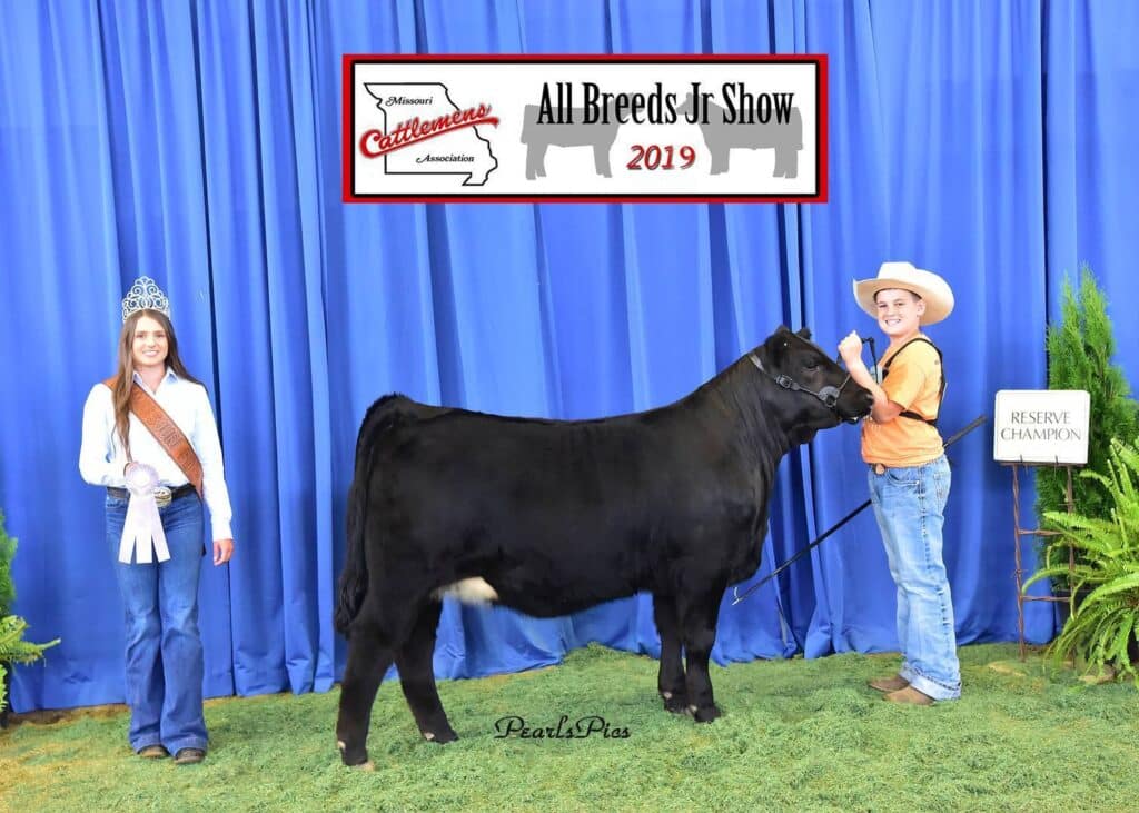 Two young women stand with a black cow at the All Breeds Jr Show 2019. One holds the cow’s lead and a plaque reading Reserve Champion; the other wears a crown and sash. A blue curtain is in the background.