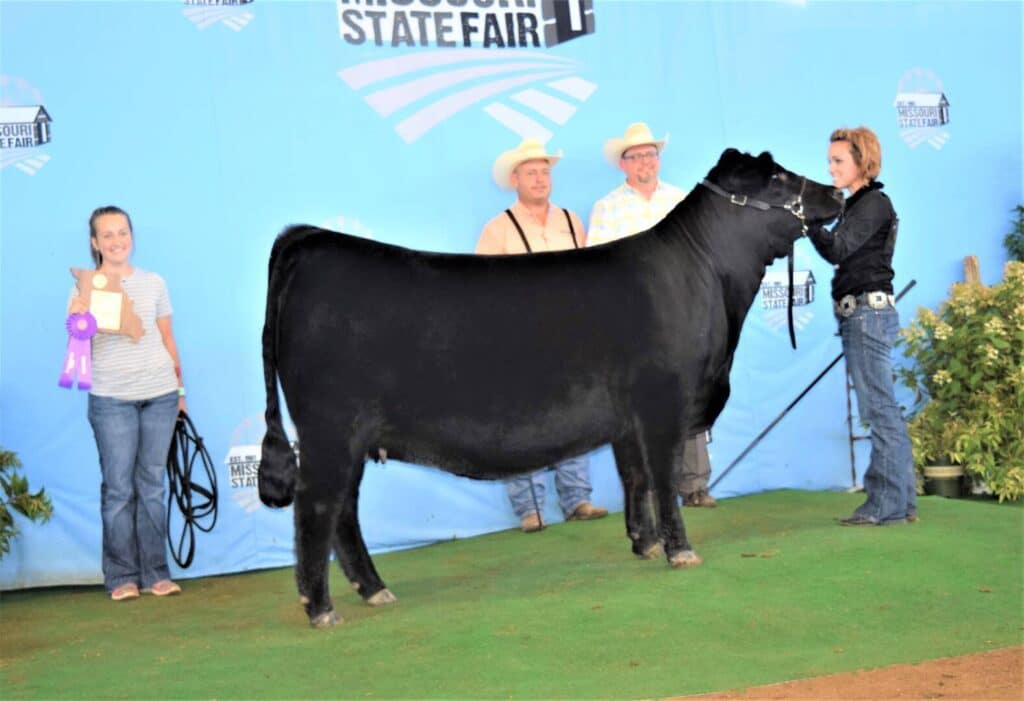 A black cow stands on green turf at the Missouri State Fair. Three people in cowboy hats pose behind the cow, while a woman holds its lead. Another woman at left smiles and displays a purple ribbon and plaque.