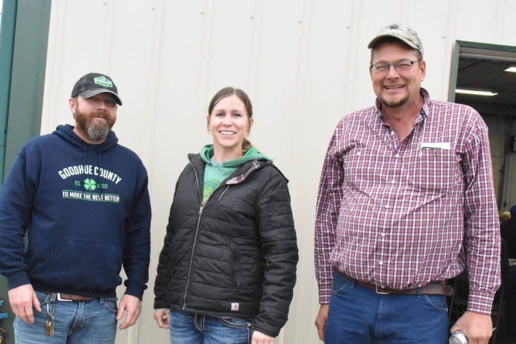 Three adults standing outdoors in front of a beige metal building, smiling. The person on the left wears a blue hoodie and cap, the center person wears a black jacket, and the right person wears glasses and a plaid shirt.